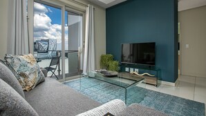 A modern living room with a sofa, a glass coffee table, a flat-screen TV, and a view of a cityscape through large windows. - Designer Apartment in The Iconic Tyrwhitt (Johannesburg)