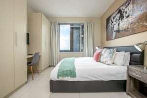A modern bedroom with a bed, a desk, a chair, a TV, and a painting on the wall. - Gorgeous Rosebank Apartment (Johannesburg)