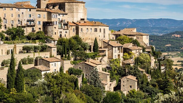 Un pueblo histórico con edificios de piedra, una torre destacada y abundante vegetación.