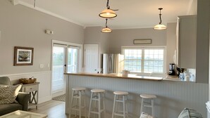 A modern kitchen with a bar area, white bar stools, and a view of the outdoors through large windows.