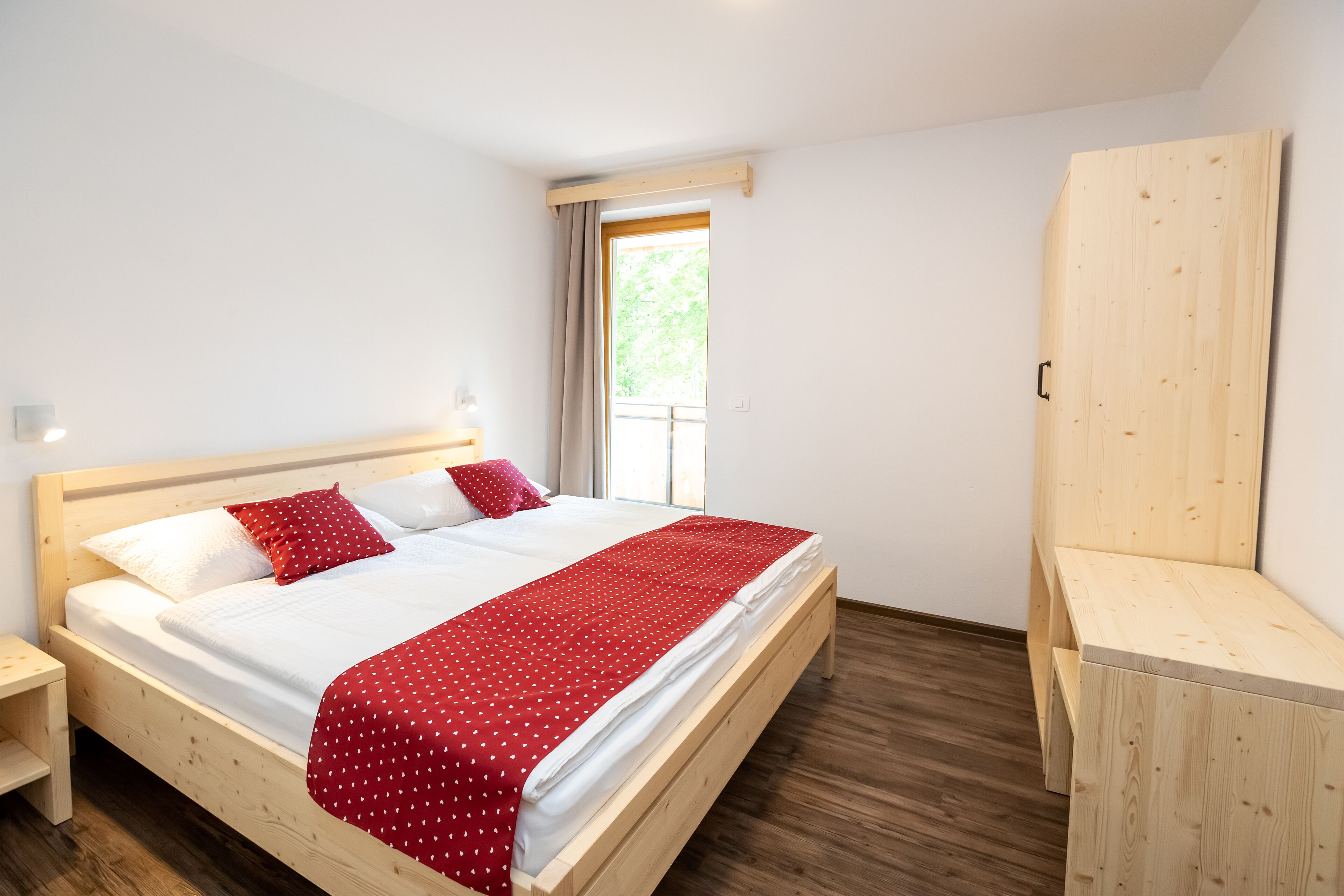 A bedroom with a wooden bed, a nightstand, a wardrobe, and a window with a view of greenery.