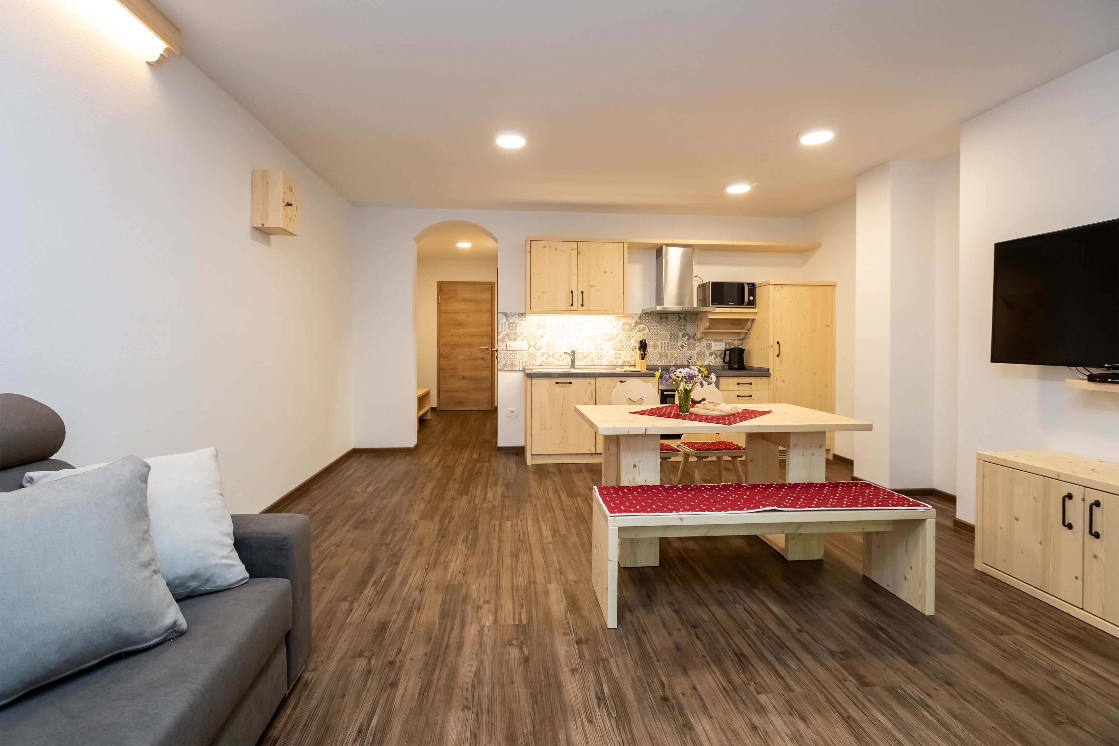 A modern living room with a grey sofa, wooden flooring, a dining table with red and white tablecloth, and a kitchen in the background.