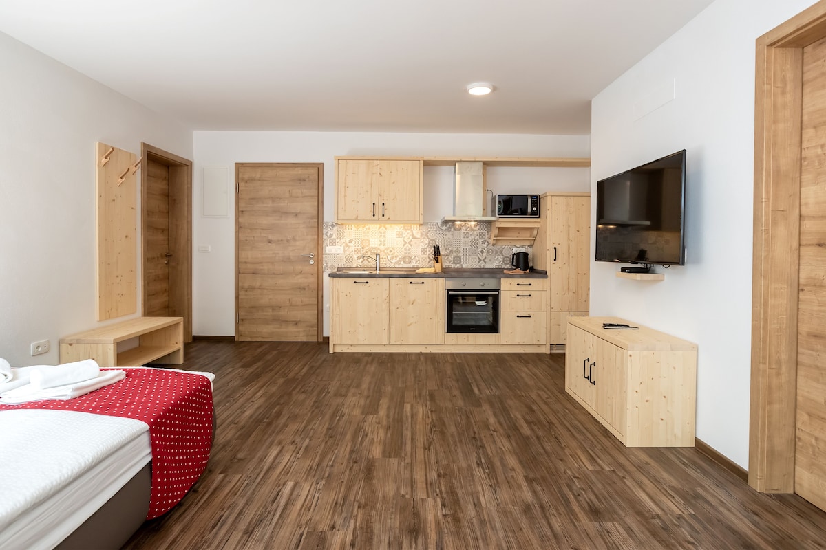 A compact hotel room with a kitchenette, a bed with a red polka dot bedspread, and a television mounted on the wall.