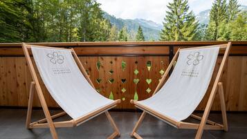 Two wooden lounge chairs with white fabric and a logo, positioned on a balcony with a view of trees and mountains.