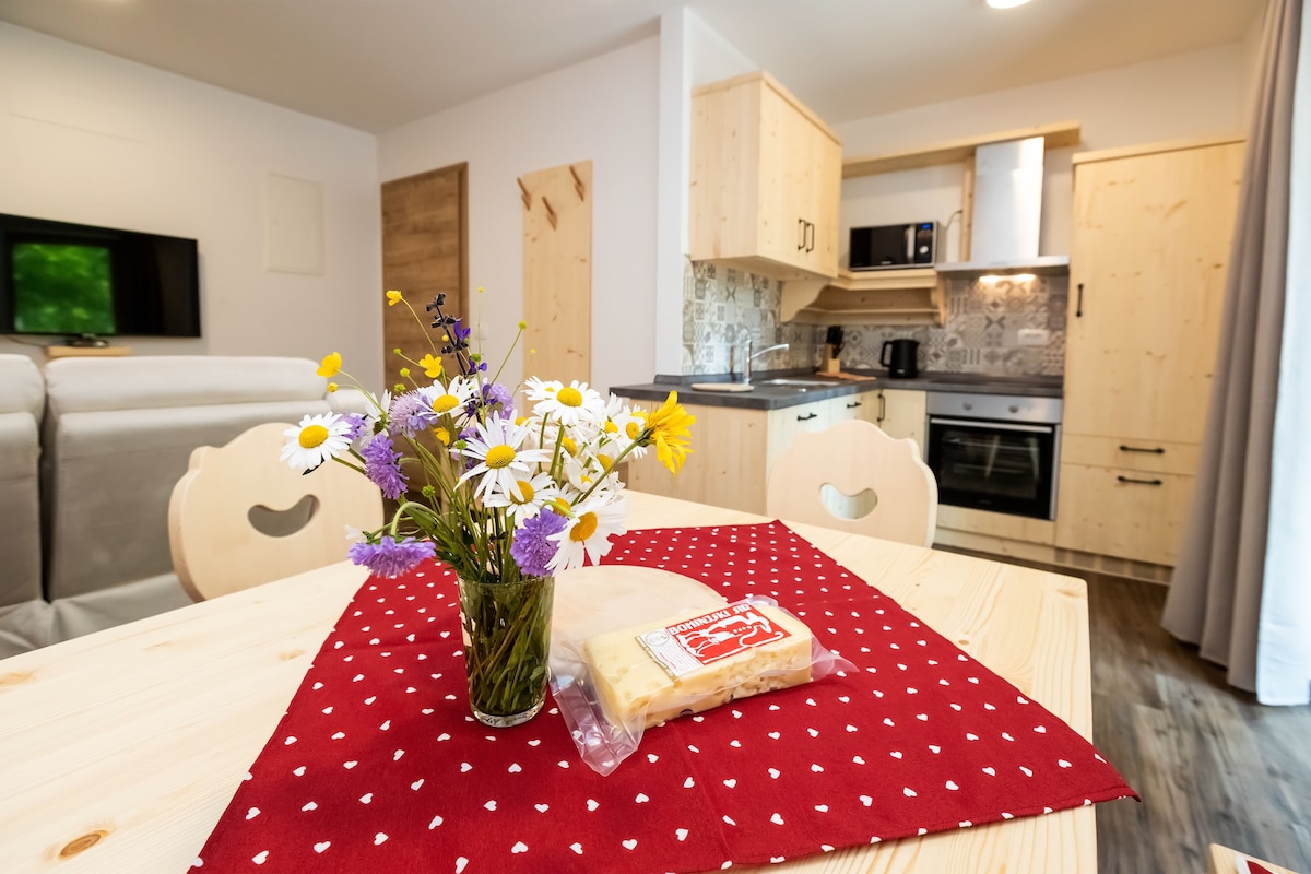 A modern living room with a sofa, a dining table with a red polka dot tablecloth, a vase with flowers, and a packaged food item.