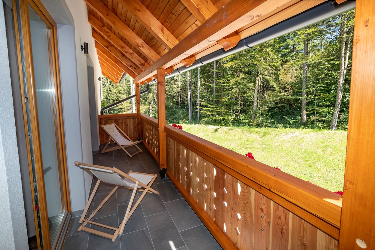 A wooden balcony with a view of a forest, a folding chair, and a door.