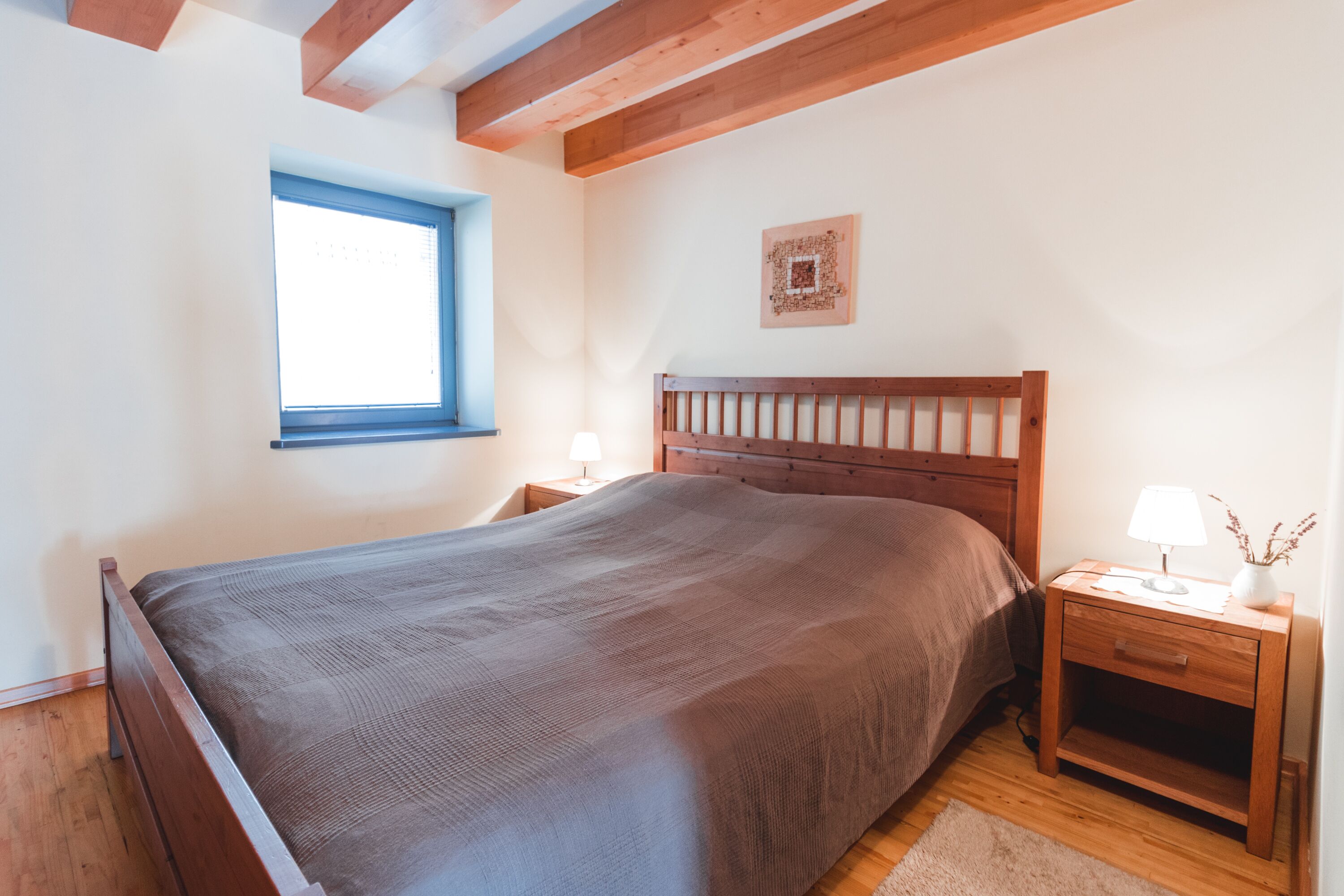 A bedroom with a wooden bed, a nightstand with a lamp, a window, and a wall-mounted artwork.