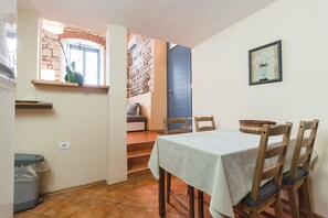 A dining area with a table set for four, wooden chairs, a trash bin, and a window with a view of a stone wall and a blue door. - Apartments Pri Mari (Piran)
