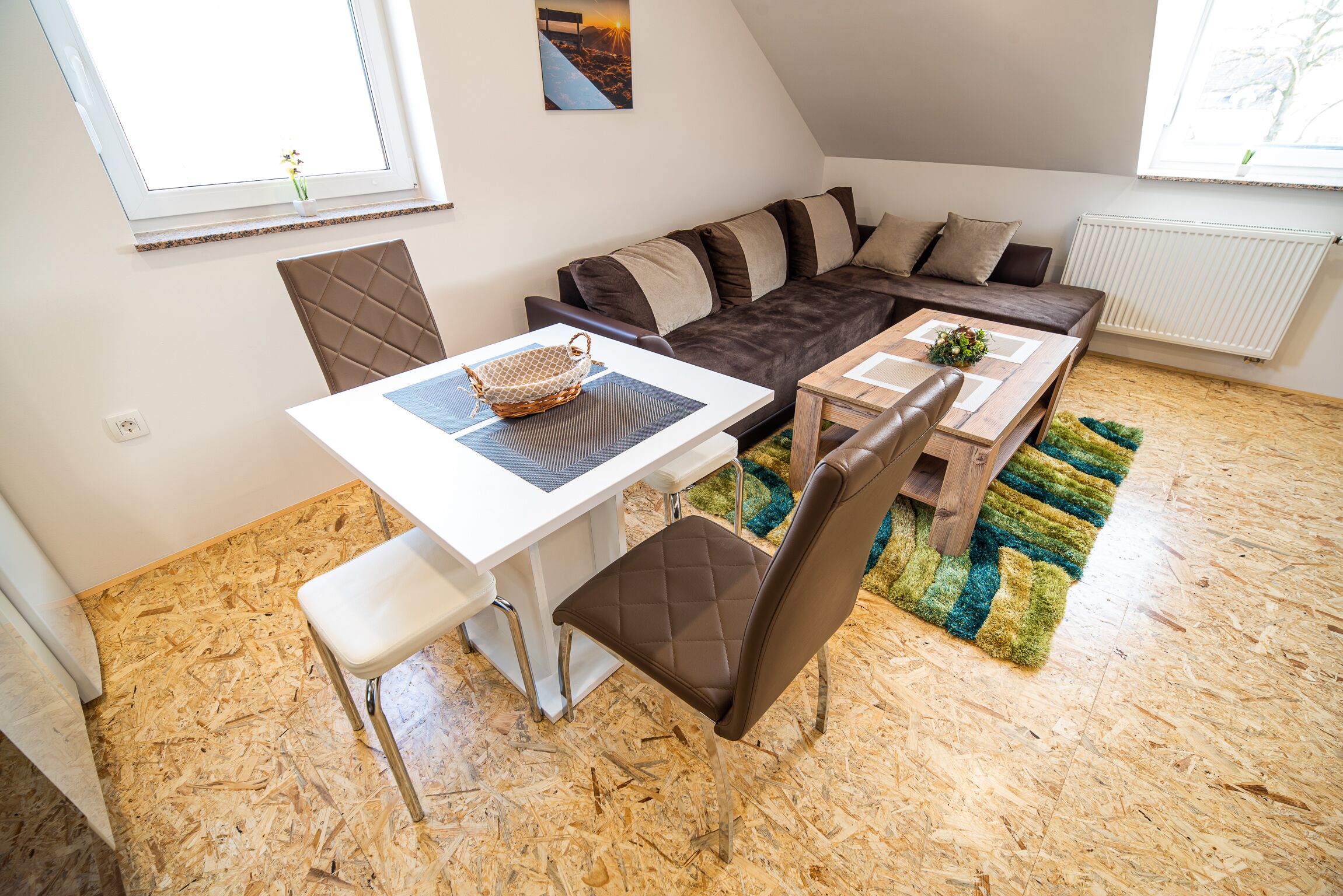 A cozy living area with a sofa, a dining table, and a chair. There is a window with a view of the outside, and a radiator on the wall.