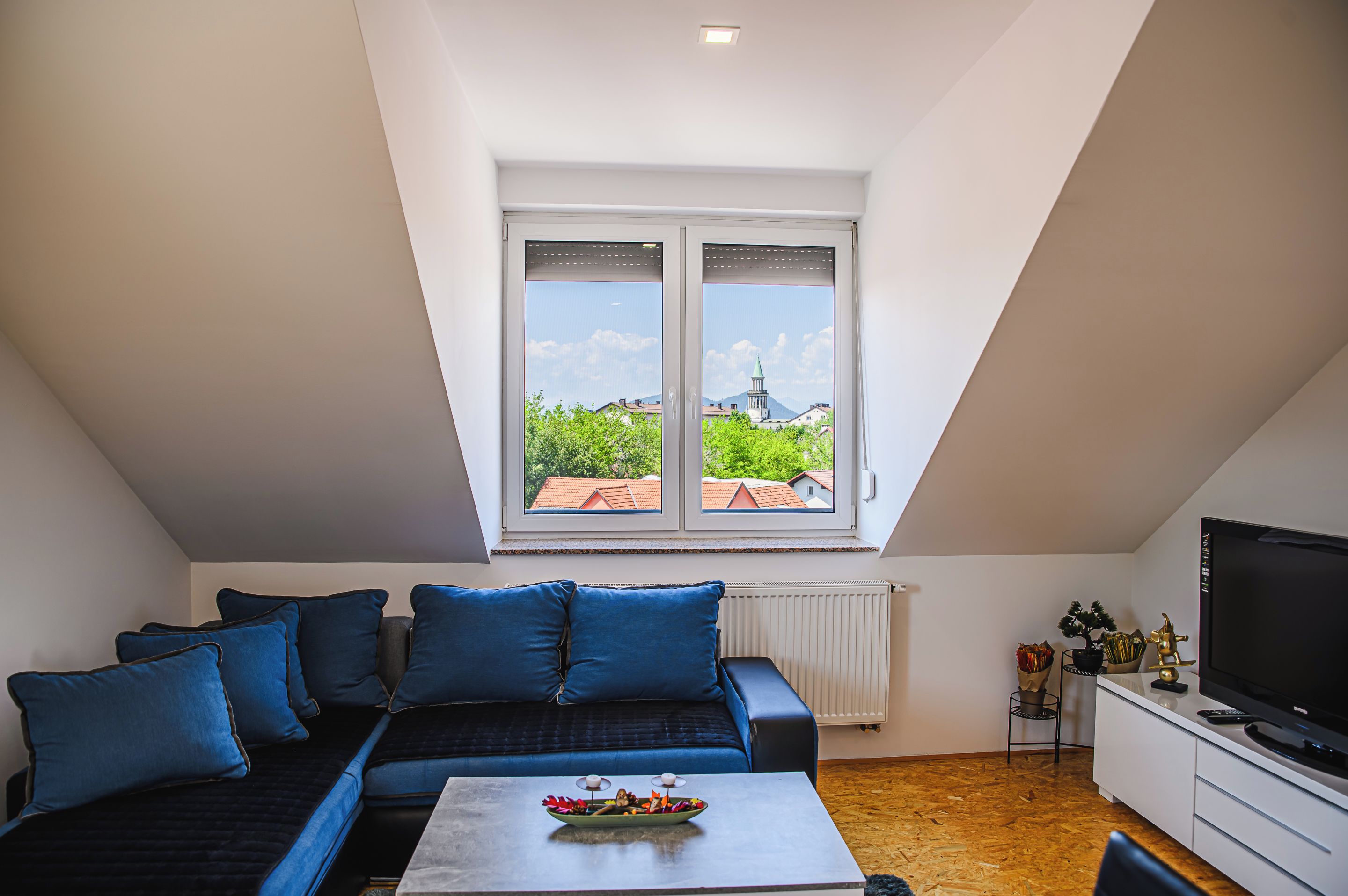 Une chambre de style loft moderne, dotée d’une grande lucarne, d’un canapé avec des coussins bleus, d’une petite table agrémentée d’objets décoratifs et d’un téléviseur posé sur un meuble.