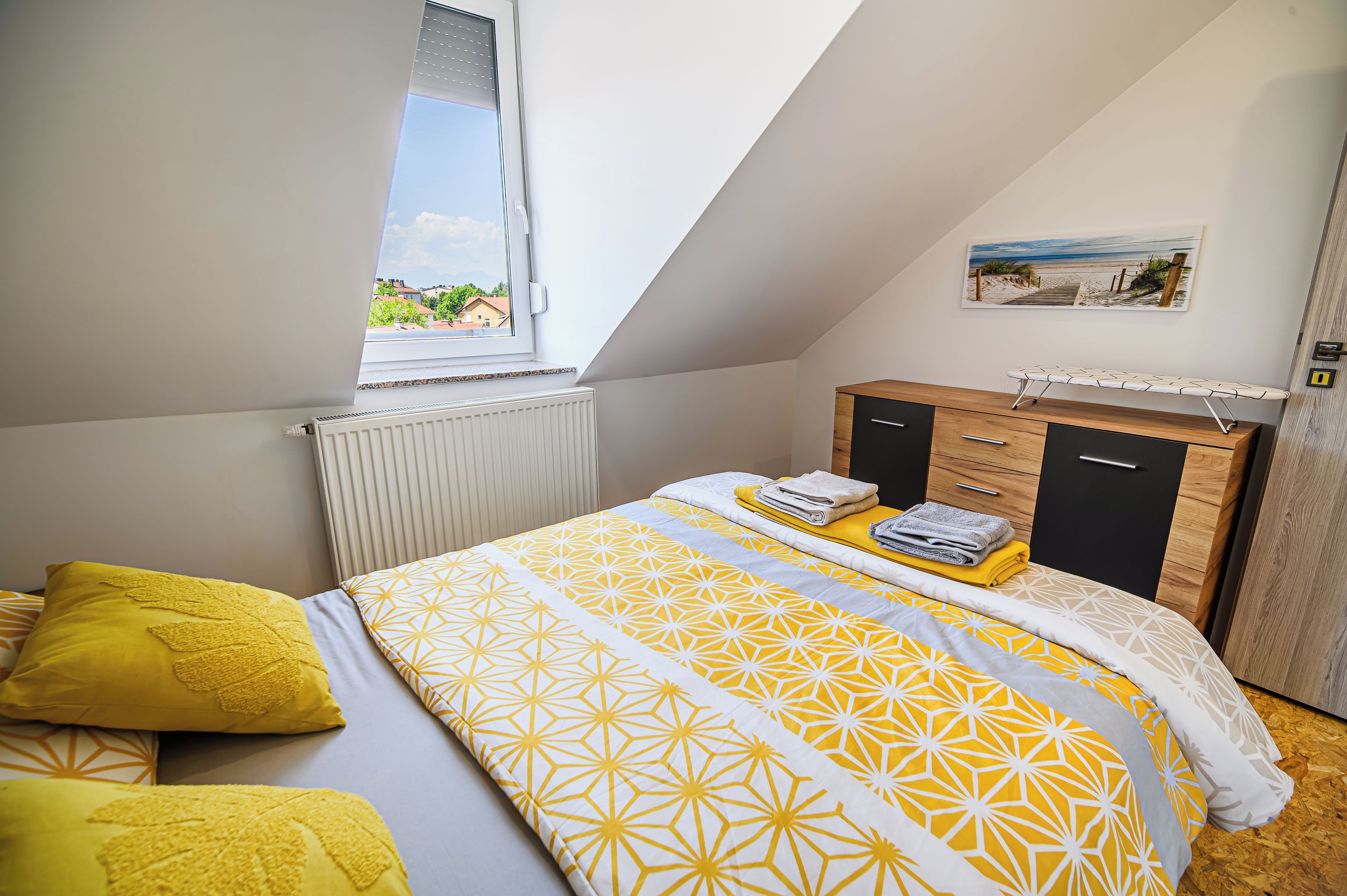 Une chambre avec un plafond en pente, un lit avec une literie à motifs, un radiateur, une commode en bois et une fenêtre donnant sur des immeubles.