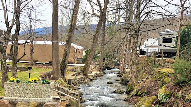 A stone bridge over a flowing stream, surrounded by trees and a distant RV campsite.