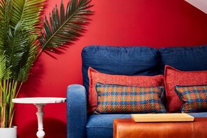A blue sofa with two patterned pillows against a red wall, a white table, and a potted plant.
