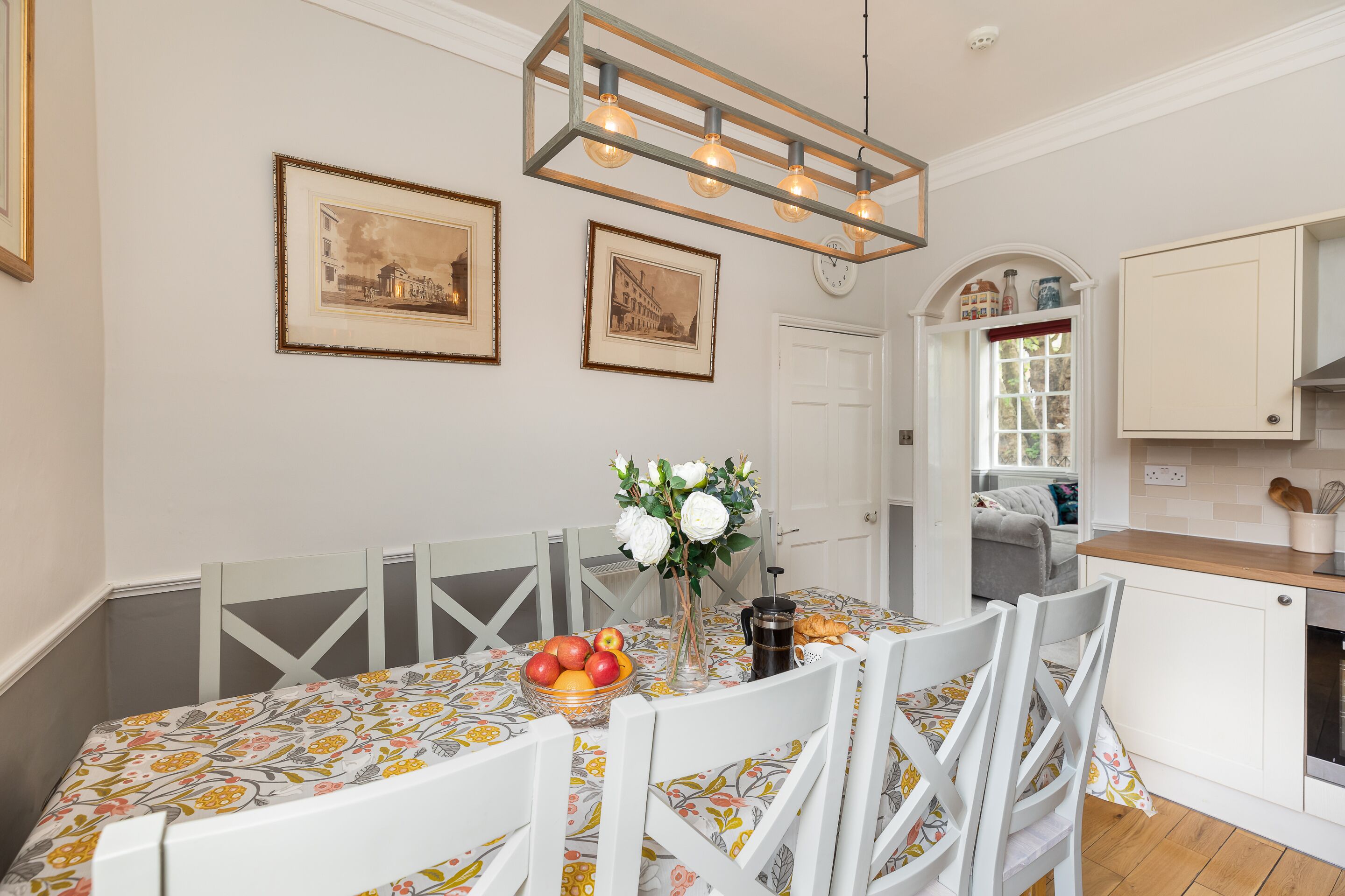 A dining area with a table set for four, surrounded by white chairs with a geometric pattern. There are two framed pictures on the wall above the table, and a vase with flowers on the table.