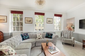 A living room with a sectional sofa, a wooden coffee table, a tufted armchair, and a large window with red valances.