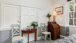 Une pièce avec deux chaises en bois, une petite table sur laquelle se trouvent des livres et un vase, une commode en bois et une fenêtre donnant sur des arbres.