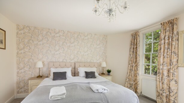 Un lit bien fait, recouvert d’une couvre-lit à rayures, avec deux lampes de chevet, une tête de lit à motifs floraux et une fenêtre avec des rideaux.