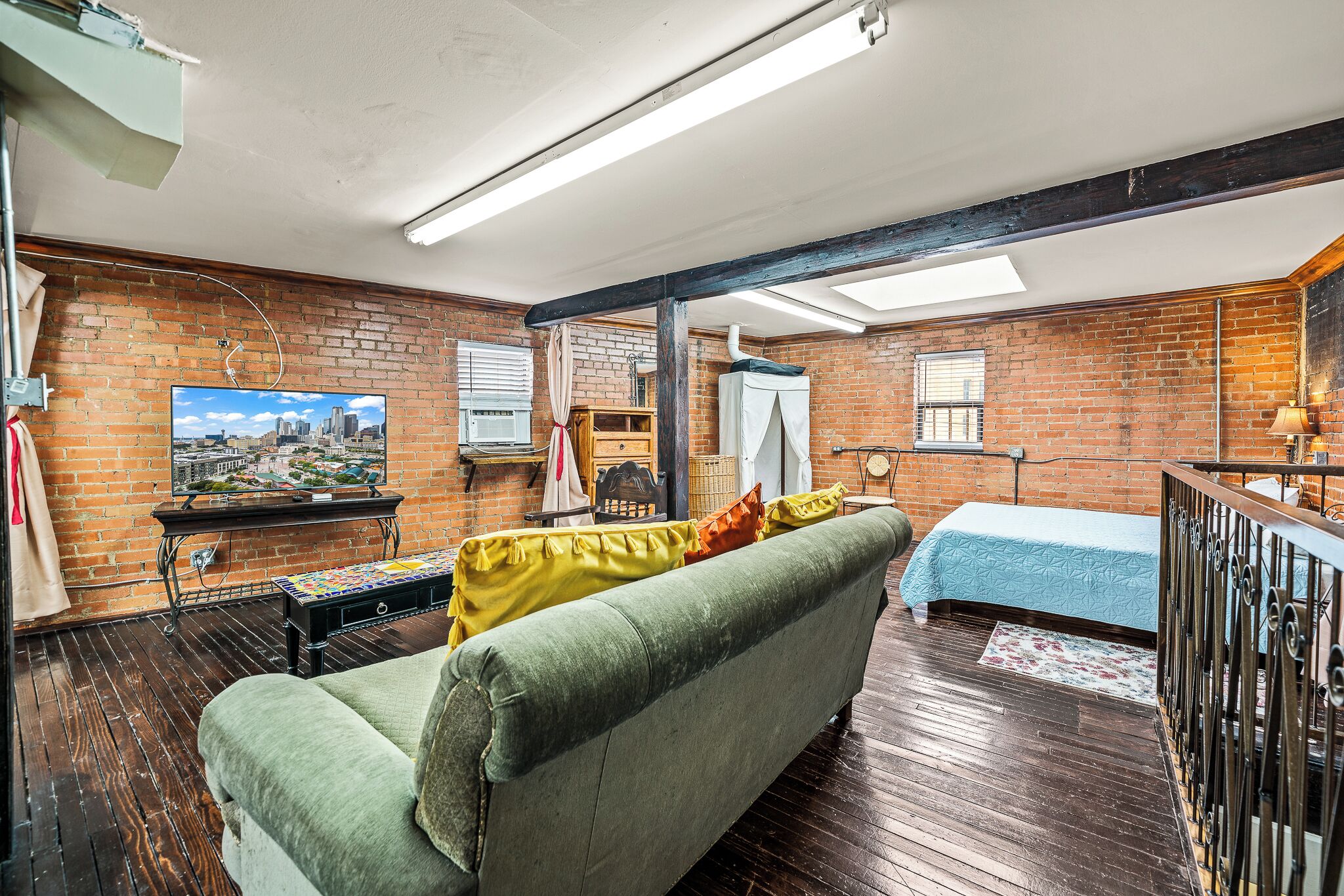 A cozy room with a green sofa, a wooden table, a TV on the brick wall, and a bed with a blue bedspread.