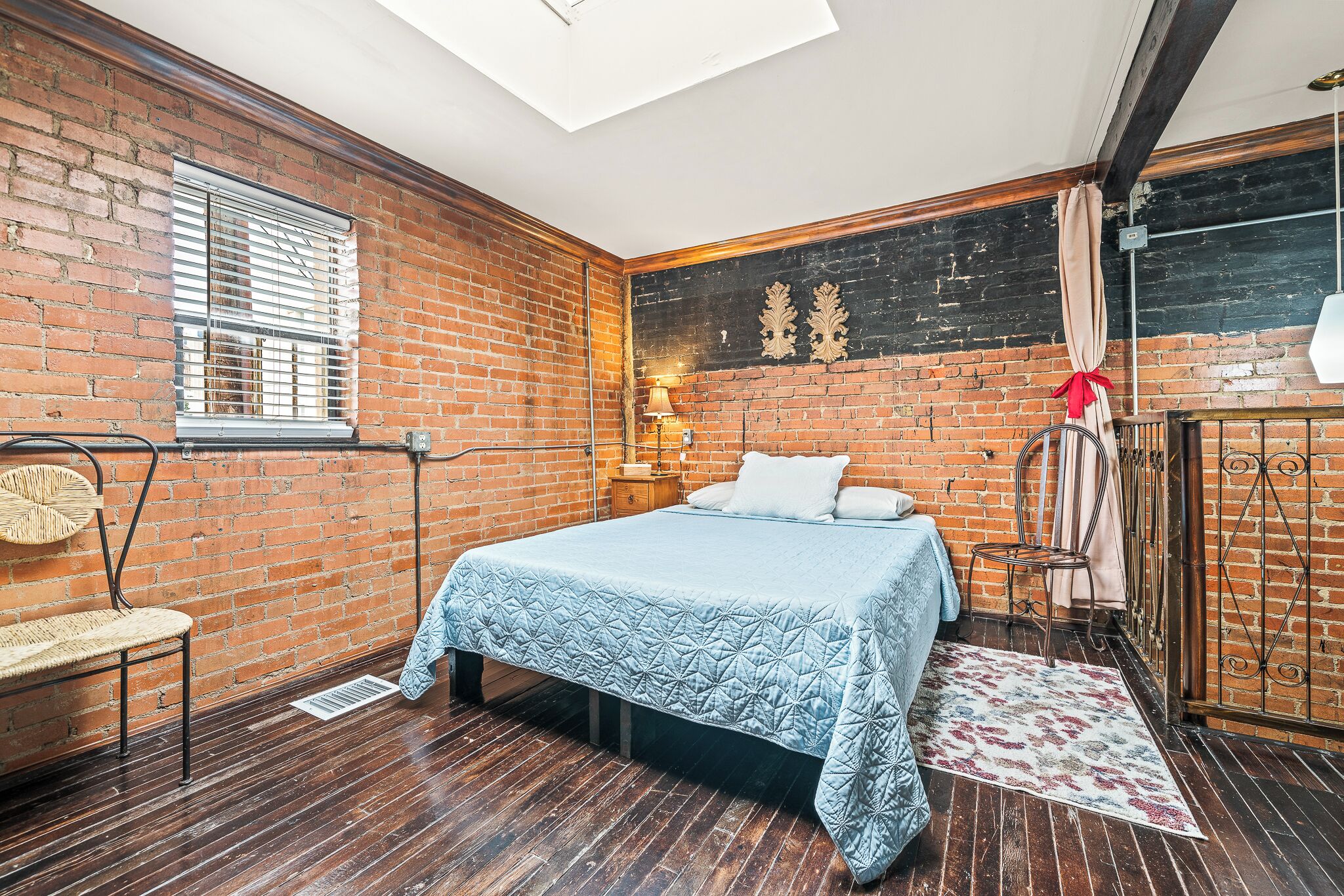 Une chambre avec un mur en briques, un lit recouvert d’une couette bleue, un plancher en bois, une fenêtre avec des stores et un puits de lumière.
