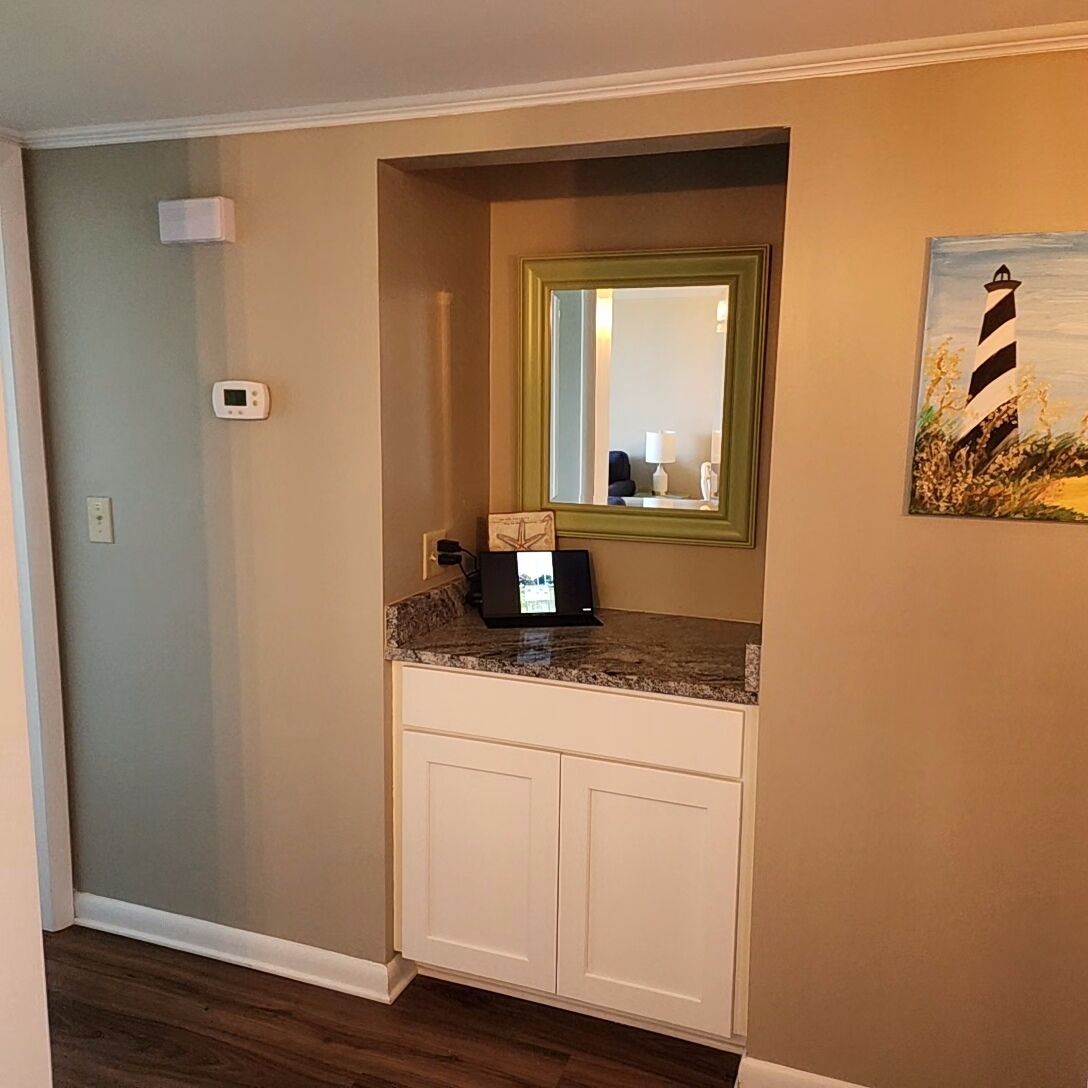 A bathroom with a granite countertop, a white cabinet, a mirror, and a painting of a lighthouse.