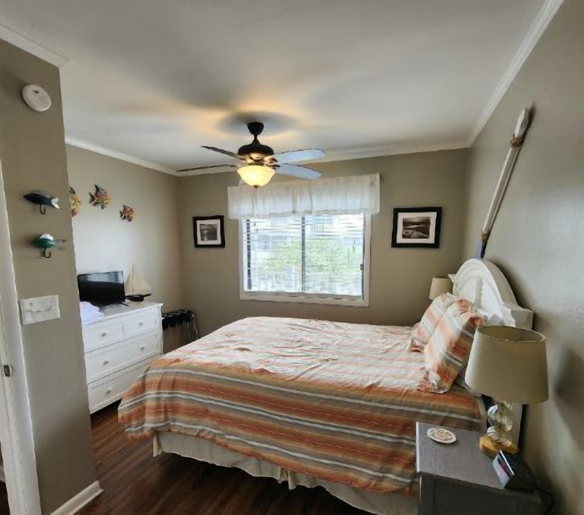 A bedroom with a bed, a nightstand, a dresser, a ceiling fan, and a window with blinds.