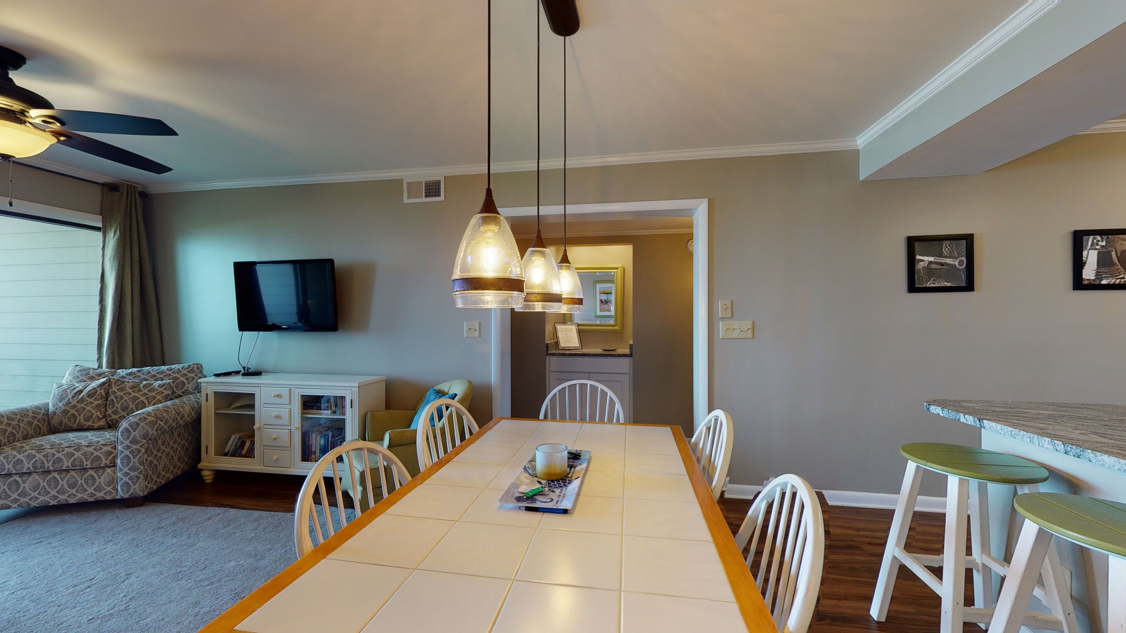 A dining area with a table, chairs, a ceiling fan, a TV, and a kitchen in the background.