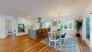 A modern kitchen with wooden flooring, a central island with bar stools, and a dining area with a large window.