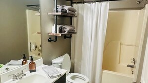 Un baño con lavamanos, inodoro y zona de ducha. Hay repisas sobre el inodoro con toallas y artículos de aseo.