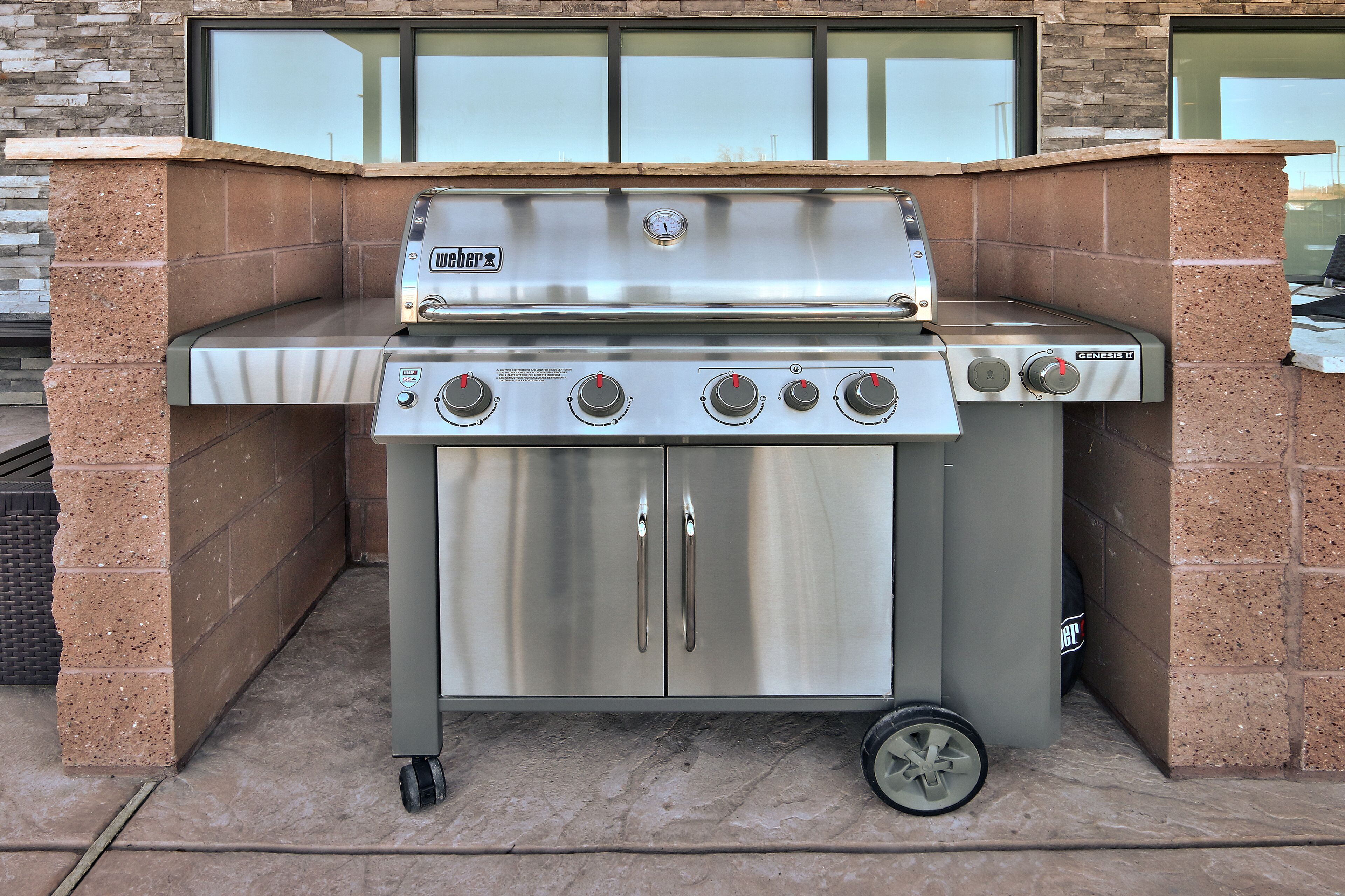 A stainless steel outdoor grill with four burners and a digital thermometer, set against a brick wall.