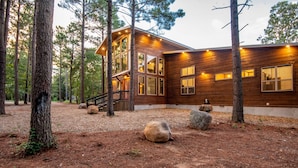 Una cabaña de madera con ventanas amplias y una terraza, rodeada de árboles y rocas.