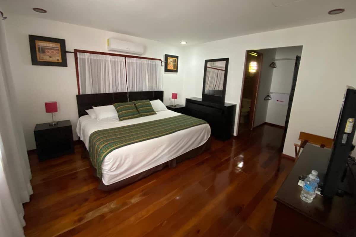 Une chambre d’hôtel avec un plancher en bois, un lit recouvert d’une couvre-lit verte et blanche, une table de chevet noire avec une lampe rouge, une commode noire et un miroir.