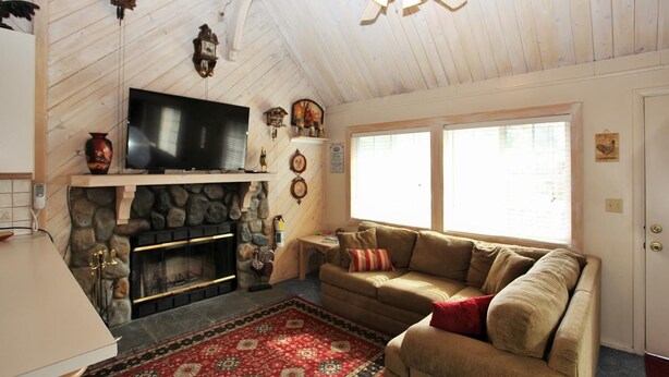 A cozy living room with a stone fireplace, a flat-screen TV, a ceiling fan, and a large window. - Cozy Cabin, Views, Walk to Bear Mountain Resort (Big Bear Lake)