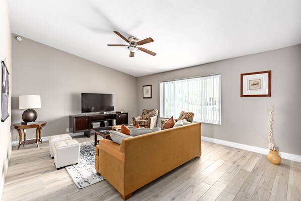 A living room with a sofa, a television, a ceiling fan, and a window with blinds.