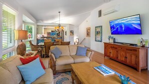 A spacious living room with a large TV, wooden furniture, and a dining area in the background. - Elegant Air-Conditioned Villa by Golf Course - Near Beaches, Shopping, Dining (Princeville)