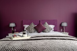A bedroom with a purple headboard, a bed with grey and white bedding, two bedside tables with lamps, and a bedside table with books and a coffee pot. - Cart House 4 bedrooms - sleeps 9 guests  in 4 bedrooms (Beverley)