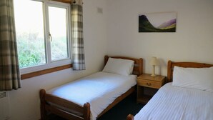 A bedroom with two single beds, a window with curtains, a bedside table with a lamp, and a painting on the wall.