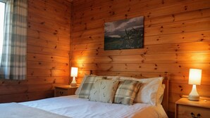 Un dormitorio de cabaña de madera con una cama, dos lámparas de noche, un cuadro en la pared y una ventana con cortinas.