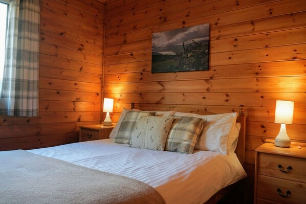 Een houten slaapkamer met een bed, twee bedlampen, een schilderij aan de muur en een raam met gordijnen.