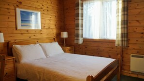 Une chambre à coucher dans un chalet en bois, avec un lit, deux tables de chevet, une petite table, une fenêtre avec des rideaux et un tableau accroché au mur.