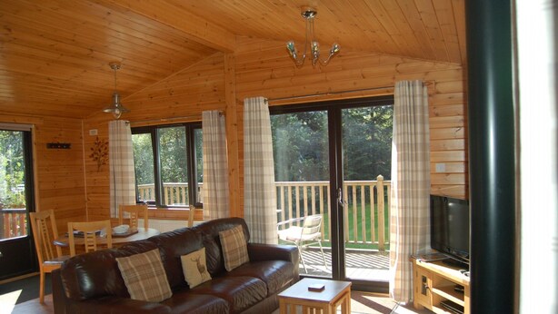 A wooden cabin interior with a leather sofa, a television, and a balcony with a wooden railing.