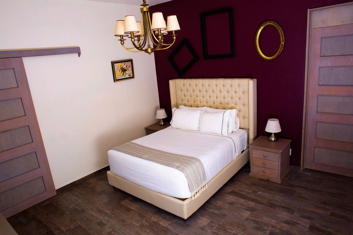 A bedroom with a large bed, a nightstand, a lamp, a chandelier, and a mirror on the wall.