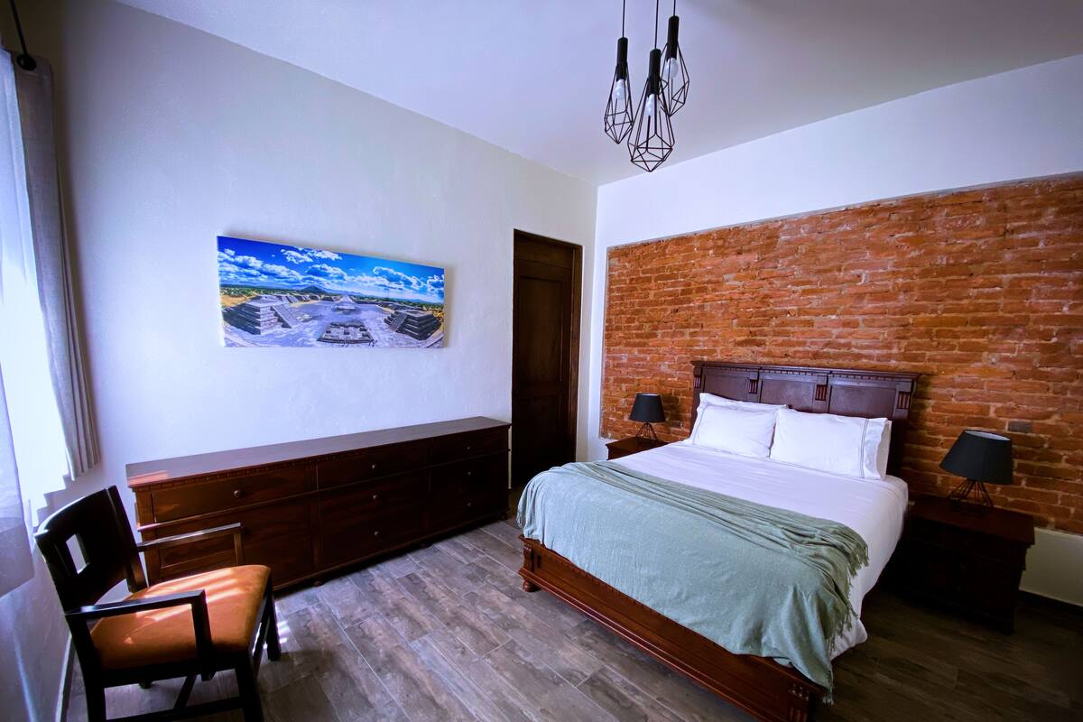 A bedroom with a brick wall, a bed, a dresser, a chair, and a painting on the wall.