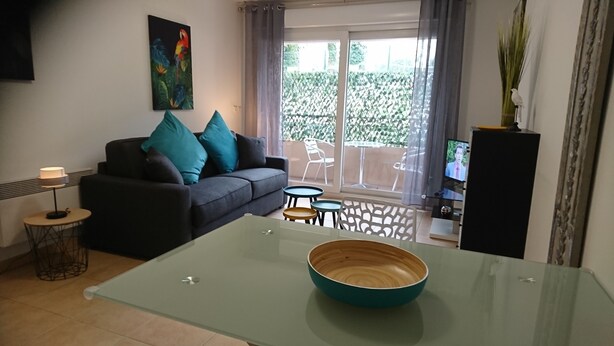 Ein Wohnzimmer mit Sofa, Couchtisch, Fernseher und einem Balkon mit Blick auf Grünflächen.