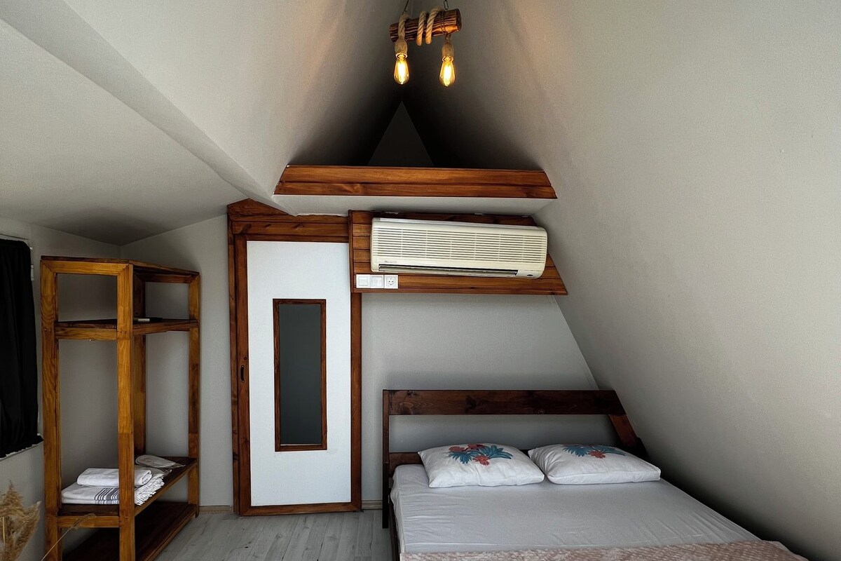 A bedroom with a sloped ceiling, a bed, a wooden shelf unit, a wall-mounted air conditioner, and a hanging light fixture.