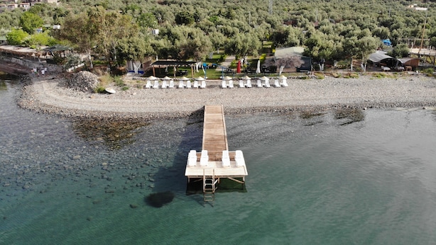 Un muelle de madera se adentra en un lago tranquilo, bordeado por una costa rocosa y una zona boscosa, con edificios al fondo.