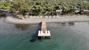 A wooden pier extending into a clear lake, surrounded by a rocky shoreline and a forested area with buildings in the background. - Concept Bungalow (Ayvacik)