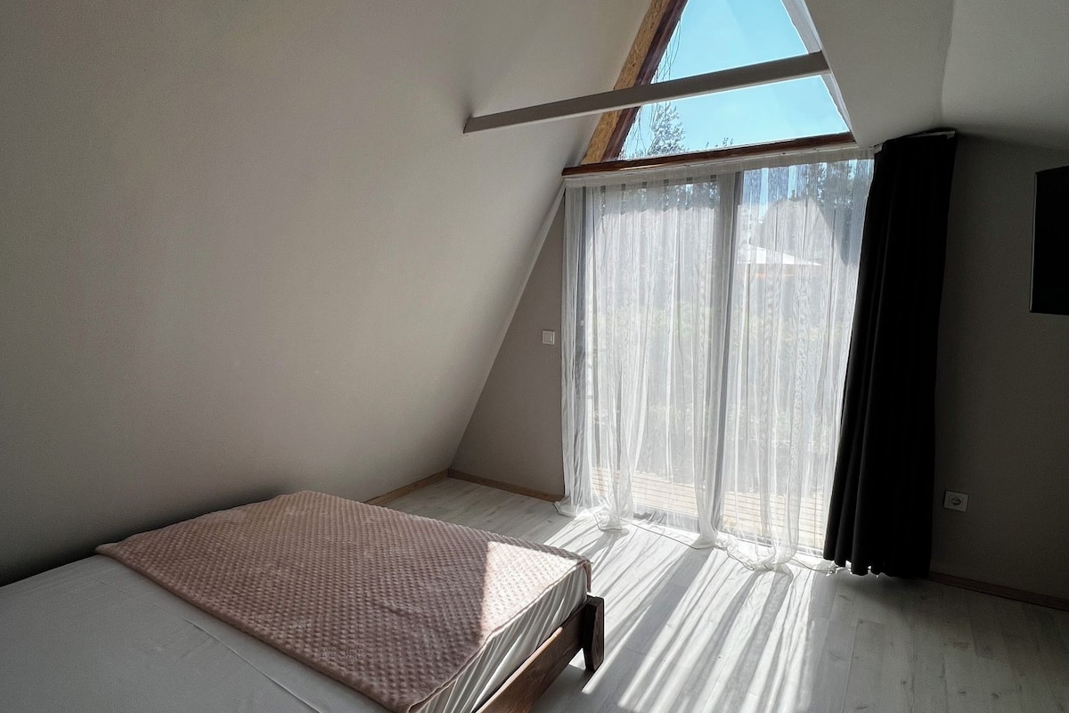 A bedroom with a sloped ceiling, a bed, a window with sheer curtains, and a wooden floor.