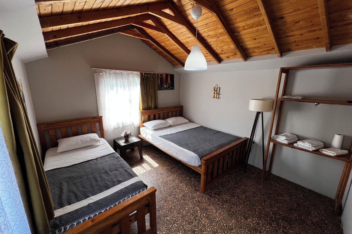 A room with two wooden beds, a small table with a lamp, a shelf with towels, and a wooden ceiling.