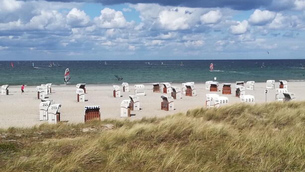 Une plage avec des cabanes de plage numérotées et une vue sur la mer, où l’on peut apercevoir des voiliers.