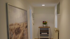 Un couloir doté d’un plancher en bois, une grande peinture sur le mur de gauche, une petite table sur laquelle est posé un vase de fleurs, et un radiateur sur le mur de droite.